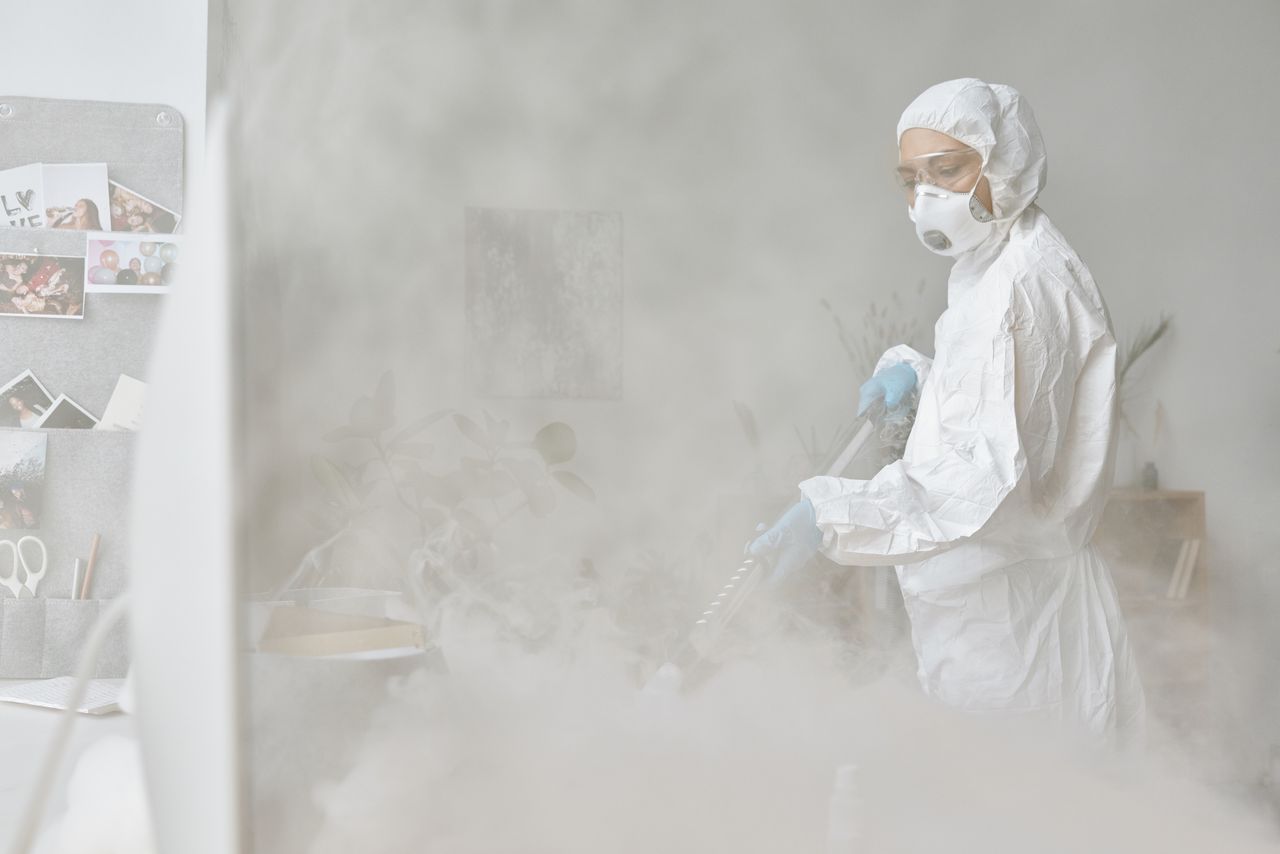 A woman in a protective suit sanitizes an indoor area using fogging equipment.